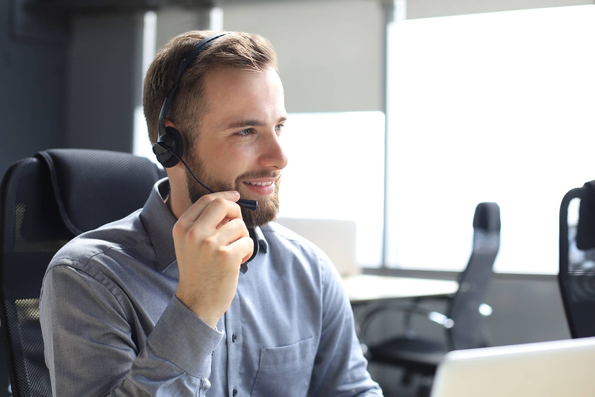 Agente de atención al cliente con auriculares y micrófono, sonriendo frente al ordenador en una oficina